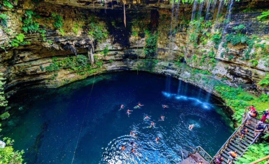 "Large cenote in Tulum with crystal-clear blue water, surrounded by lush vegetation, and visitors swimming and enjoying the natural beauty.